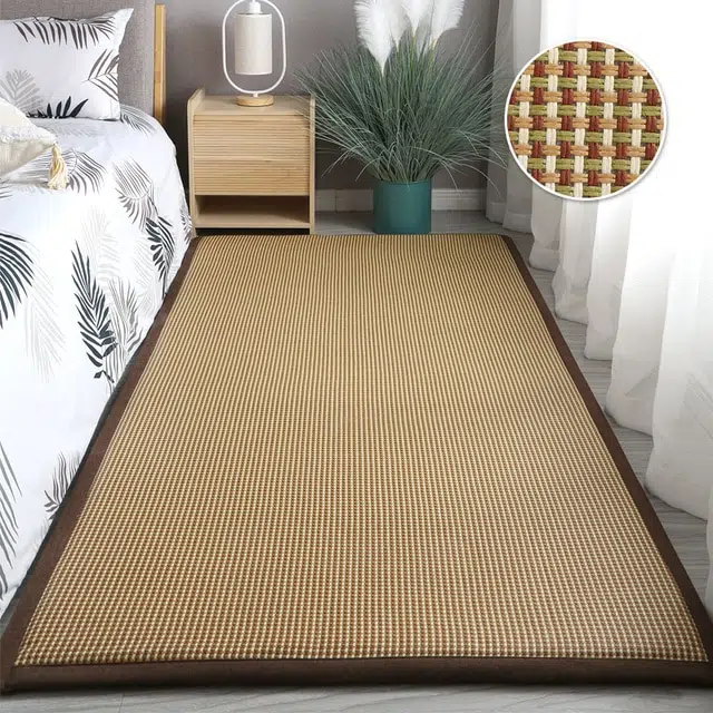 Tapis rectangulaire en rotin marron et vert. Il est posé à côté d'un lit, on voit également une table de chevet et un rideau blanc.