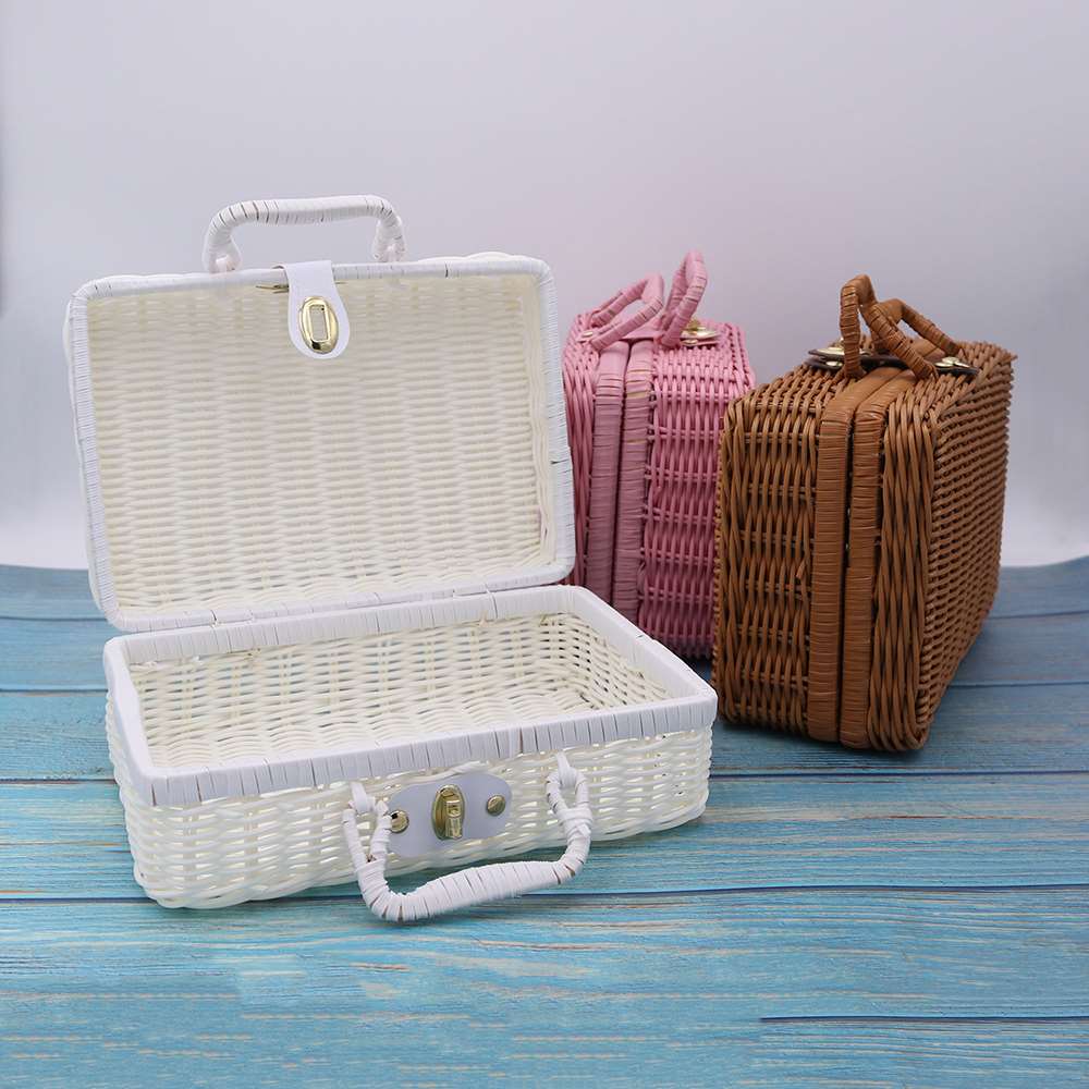 Trois valises en rotin sont posées sur du bois bleu. elles sont blanches, rose et marron. La valisette blanche en rotin est ouverte.