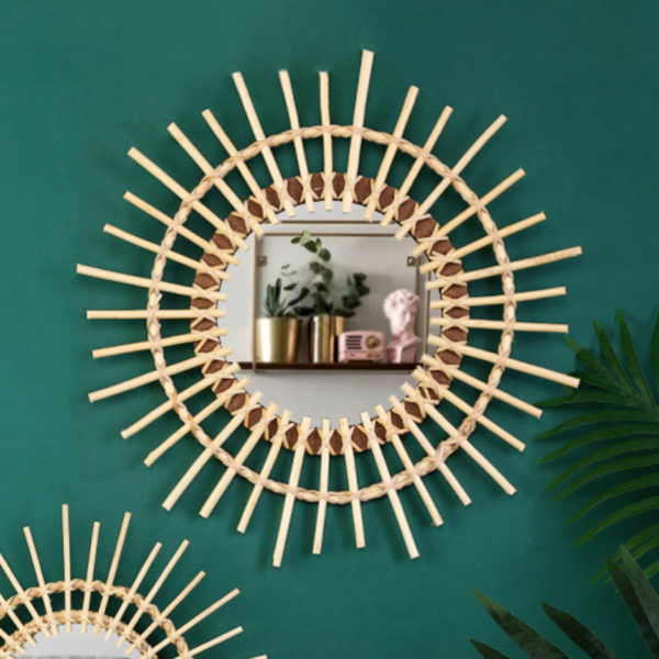Miroir circulaire en rotin en forme de soleil sur un mur vert. On voit une plante à droite et une étagère dans le reflet du miroir.