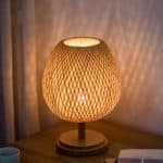 Cette lampe de chevet est ronde et en rotin. Elle est placée sur une table de chevet en bois. le socle de la lampe est en bois.