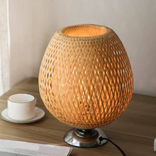 Cette lampe en rotin est ronde et a un socle en métal. Elle est posée sur une table en bois avec une tasse blanche et un journal.