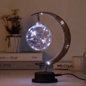 Cette lampe est argentée et en forme de lune . Elle est placée sur un meuble avec des livres en fond.