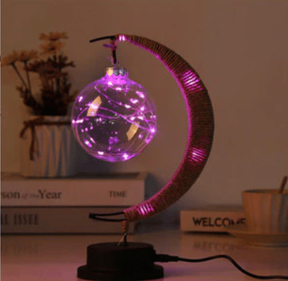 Cette lampe est rose et en forme de lune . Elle est placée sur un meuble avec des livre en fond.