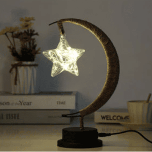 Cette lampe est en forme de lune avec une ampoule en étoile . elle est posée sur un meuble avec des livres en fond.