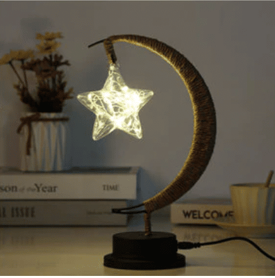 Cette lampe est en forme de lune avec une ampoule en étoile . elle est posée sur un meuble avec des livres en fond.