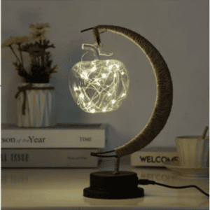 Cette lampe est en forme de lune avec une ampoule pomme . elle est posée sur un meuble avec des livres en fond.