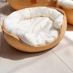 Panier circulaire en rotin beige avec un coussin blanc style traversin et un coussin plat pour créer le lit, pour animaux. Il est posé sur un carrelage blanc.