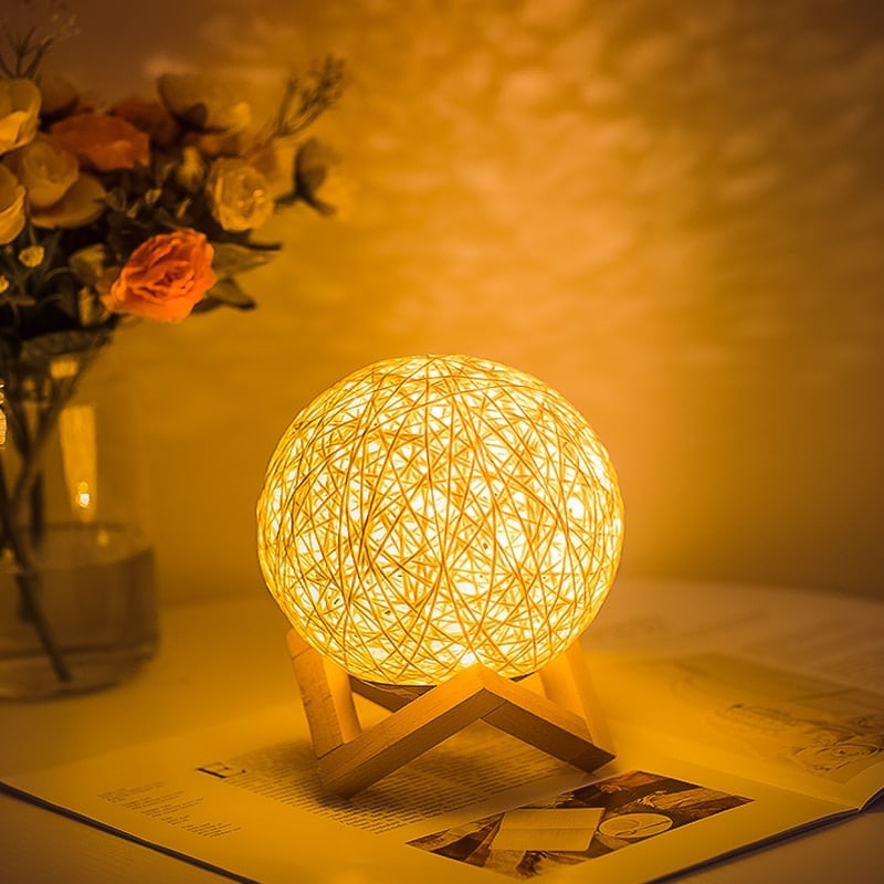 Lampe de chevet en rotin en forme de boule sur un pied en bois sur une able de nuit avec un bouquet de fleur dans un vase à gauche de la lampe