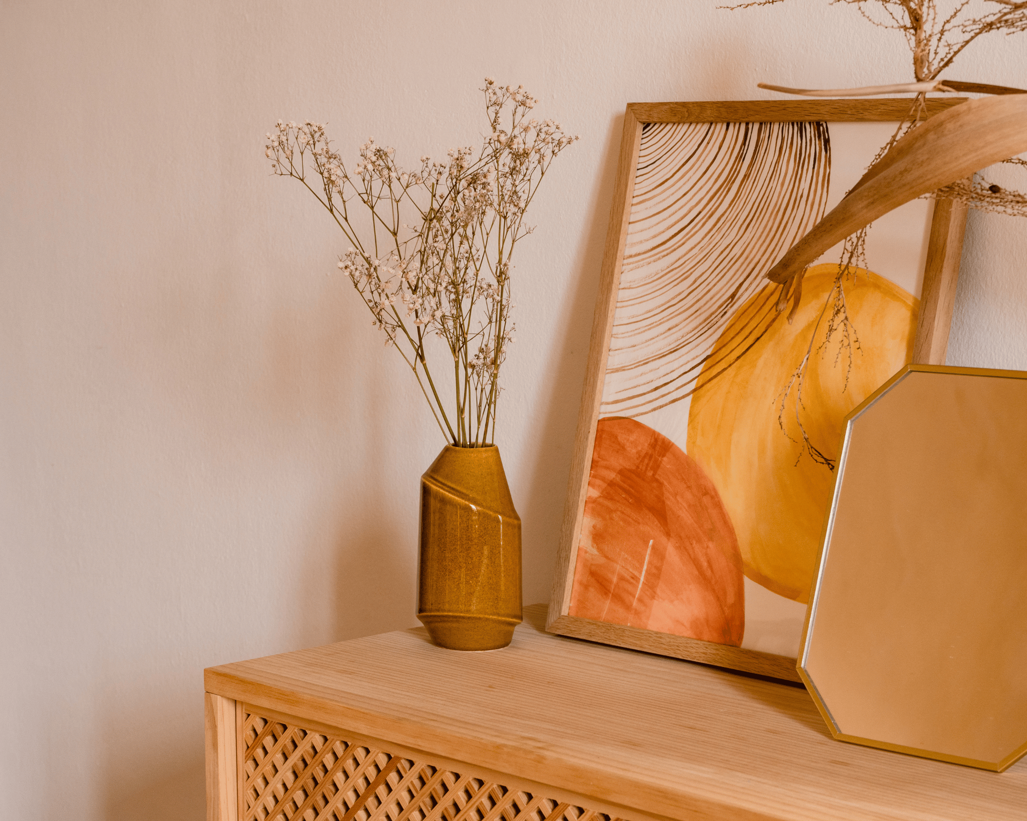 meuble en rotin sur lequel se trouve u vase en rose avec desplantes dedans , un cadre avec un tableau coloré par des formes arrondies et un cadre orange