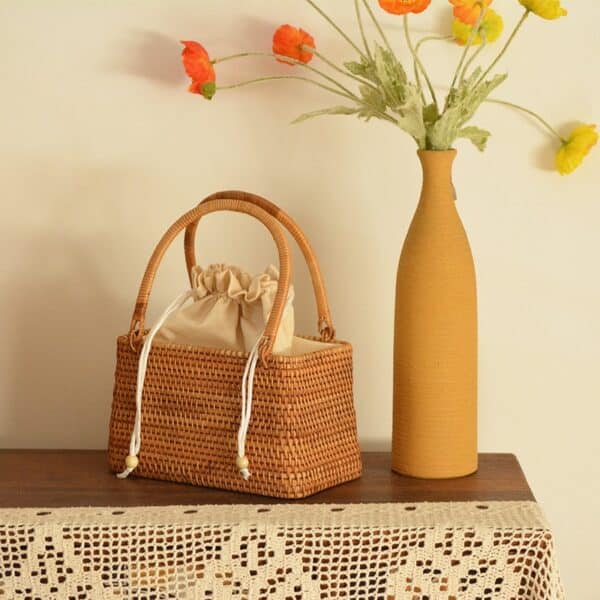 Petit sac en rotin rectangulaire avec des hanses et un sac en coton dedans avec fermeture à lanière sur une table avec un vase à droite avec des fleurs dedans