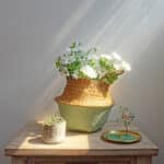 Sur une table, un panier en rotin bicolore , dont le haut est couluer naturelle et le bas vert clair, contient des fleurs , devant sont posés un bibelot et une petite sculpture en pierres précieuse assortie