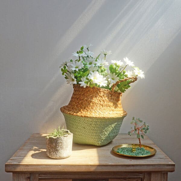 Sur une table, un panier en rotin bicolore , dont le haut est couluer naturelle et le bas vert clair, contient des fleurs , devant sont posés un bibelot et une petite sculpture en pierres précieuse assortie