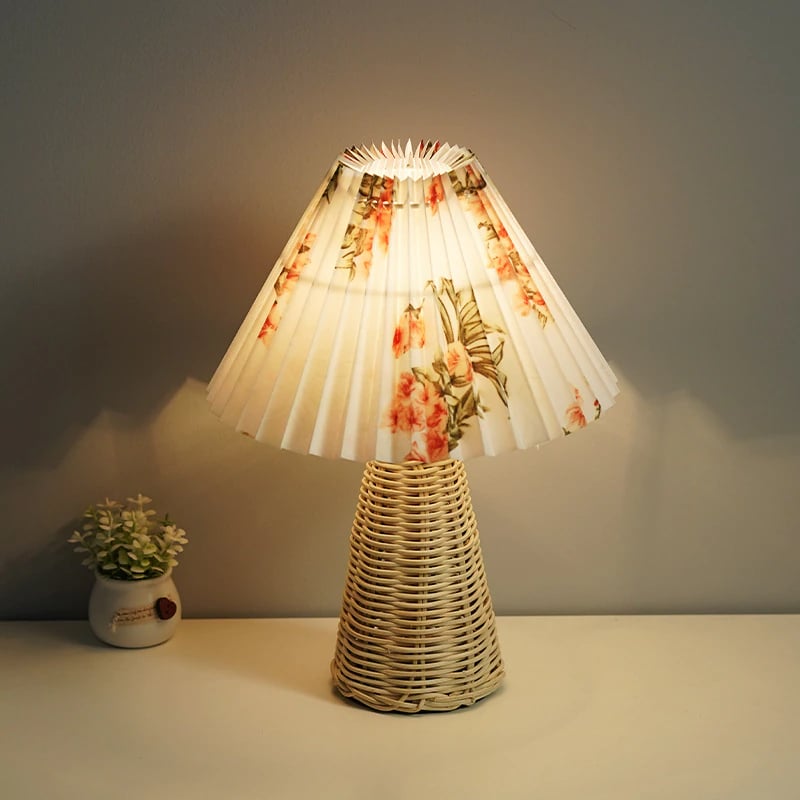 Posée sur une table , une lampe avec la base en forme de cône en rotin, et l'abat-jour plissé blanc avec des motifs fleuris