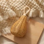 Hochet pour bébé en rotin naturel de couleur marron et la anse beige, posé sur un support en bois et près d'un tissé tricoté beige à franges