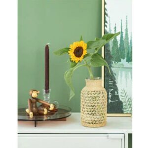 Vase en verre avec une décoration en rotin tressé avec un tournesol dedans, sur une table blanche devant un tableau dans les tons vert et près de bibelots