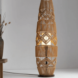 Lampadaire Osier à LED Luxueux Tricoté à la Main allumé sur fond gris avec une plante dans un pot posée sur un meuble à gauche