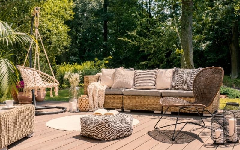 Salon d'extérieur en rotin, avec une chaise suspendue, un canapé avec plusieurs coussins, une chaise, un pouf, un tapis sur une terrasse en bois. On voit des arbres derrière.