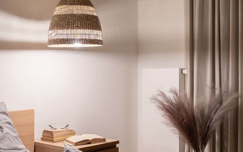 Comment installer une suspension en luminaire ? Suspension en rotin, dans une pièce blanche avec des décorations telles que plumeau et rideau et livre sur petite table de nuit.