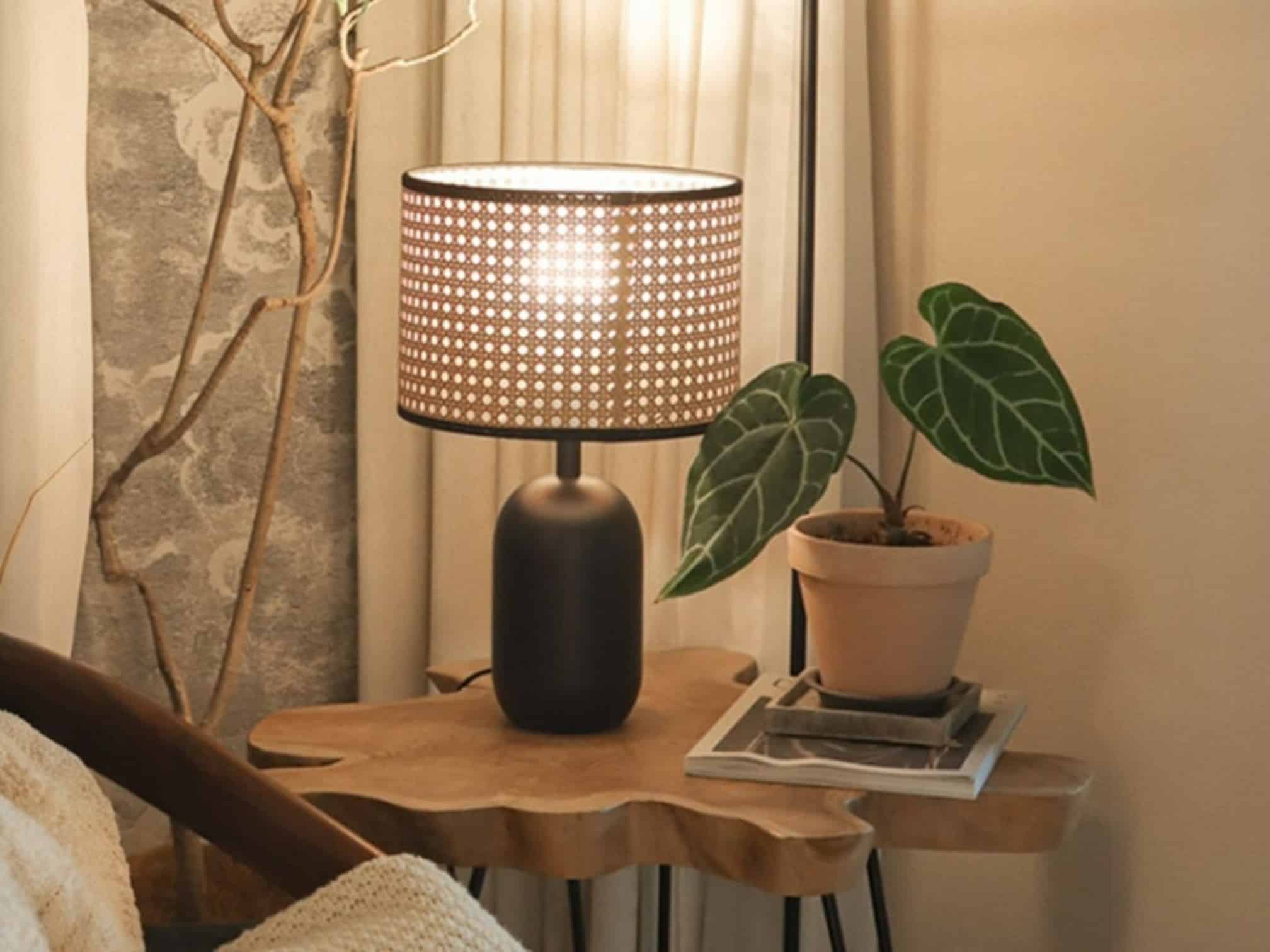 Où et comment placer une lampe en rotin dans chaque pièce de la maison ? Lampe à poser avec un abat-jour en rotin et une base en céramique noire, installée sur une table d'appoint avec un plateau en gros morceau de bois, une plante verte à côté, le tout près d'un fauteil.